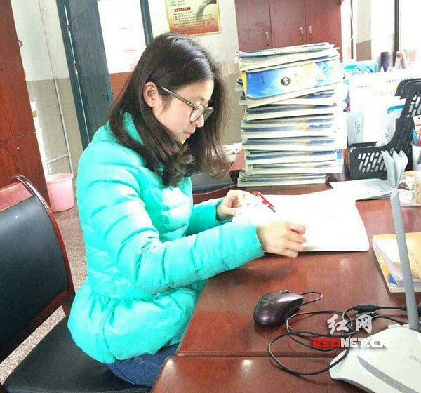 赵丽丽老师在办公室批改学生作业，其左手边堆着近半米高待改作业。